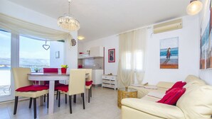 A modern living room with a beige sofa, a dining table with chairs, and a kitchen area. - Apartments Duga (Okrug)