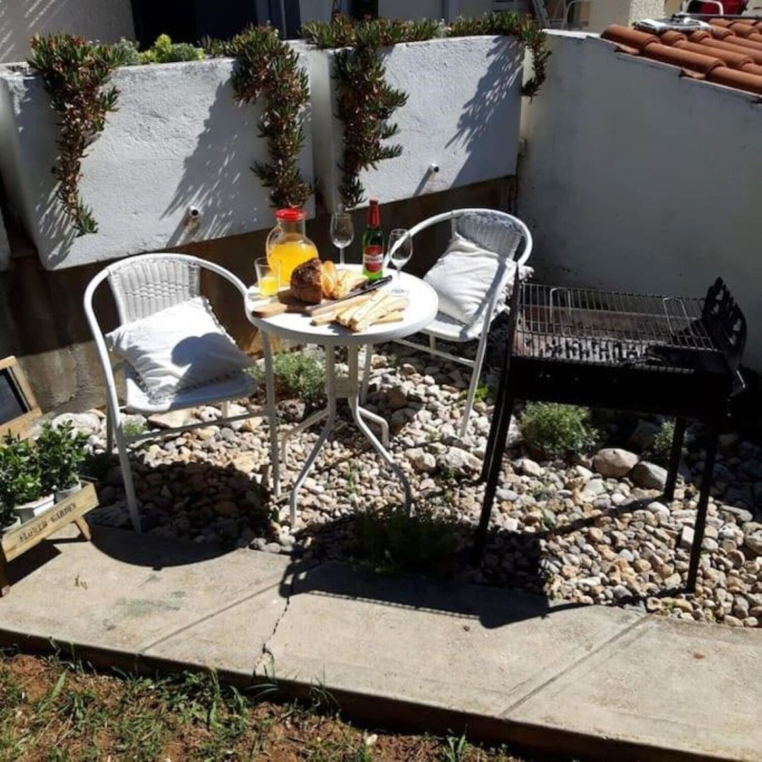 Une terrasse avec deux chaises en osier blanches, une petite table ronde sur laquelle sont disposés de la nourriture et des boissons, et un barbecue.