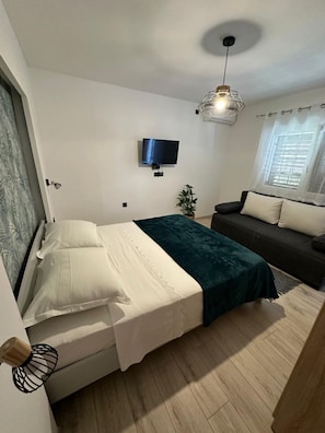 Una camera da letto compatta con un letto, una TV a muro, una piccola pianta e una finestra con persiane.