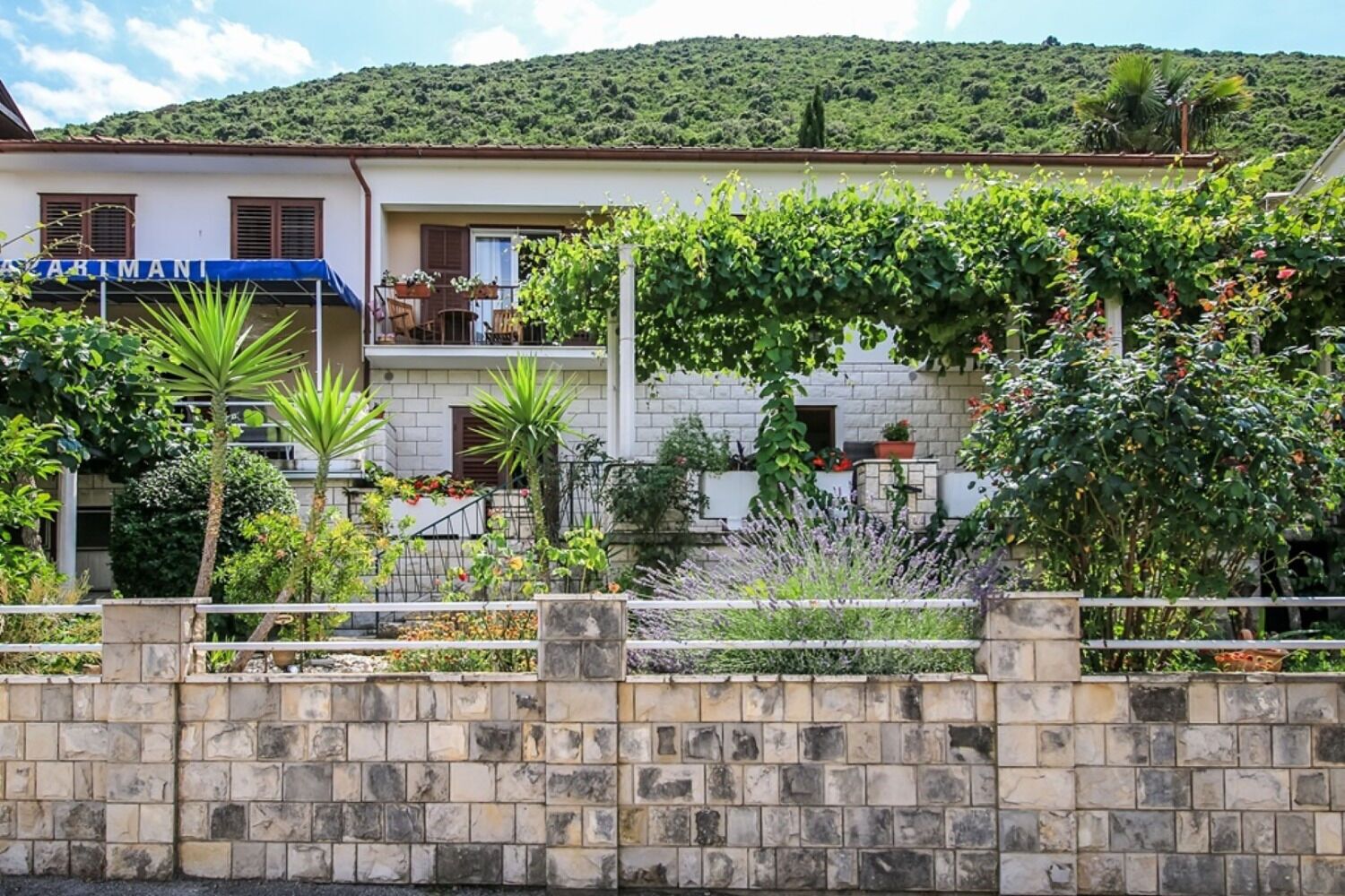 A building with a stone wall in the foreground, a balcony with plants, and a hillside in the background.