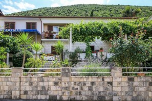A building with a stone wall in the foreground, a balcony with plants, and a hillside in the background. - Apartments Ivana (Trpanj)