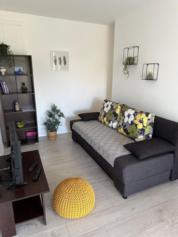 A modern living room with a grey sofa, a yellow ottoman, a TV on a wooden table, and a bookshelf with various items. - Apartments Ivana (Trpanj)