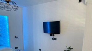 Ένα σύγχρονο υπνοδωμάτιο με ένα κρεβάτι, μια τηλεόραση τοποθετημένη στον τοίχο, ένα κομοδίνο, ένα φυτό και ένα παράθυρο με μπλε φωτισμό.