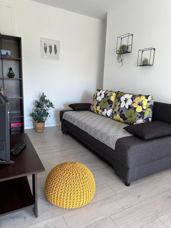 Un salon moderne avec un canapé gris, un pouf jaune et un téléviseur posé sur une table en bois.