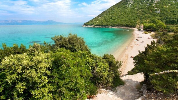 Ένα παραθαλάσσιο μονοπάτι που οδηγεί σε μια παραλία με κρυστάλλινα, γαλαζοπράσινα νερά, η οποία περιβάλλεται από πλούσια βλάστηση και έχει ως φόντο ένα επιβλητικό ορεινό τοπίο.
