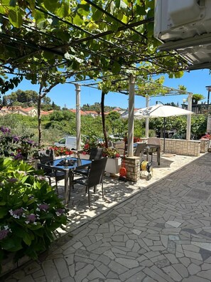 An outdoor patio with stone flooring, a table with chairs, a pergola with vines, and a white umbrella. - Apartments Ivana (Trpanj)