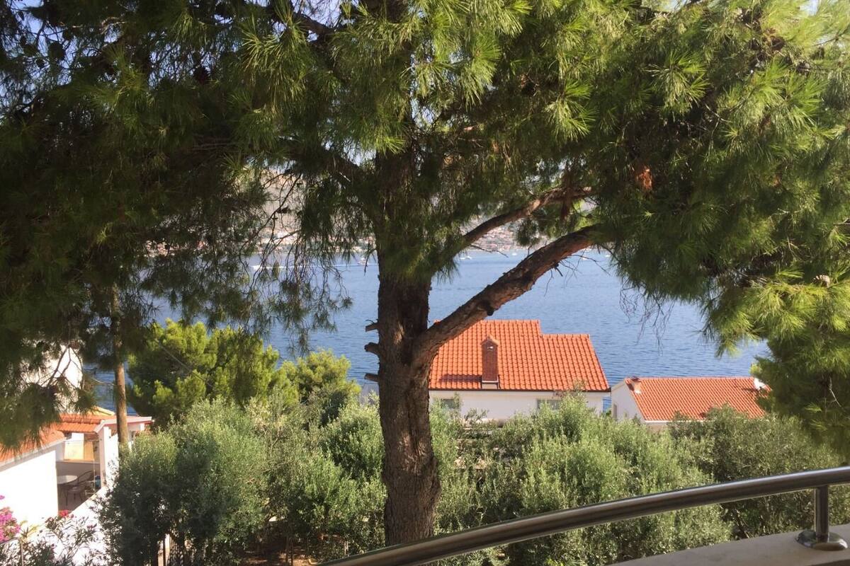 A balcony with a railing overlooking a coastal area with a red-roofed building and a tree in the foreground.