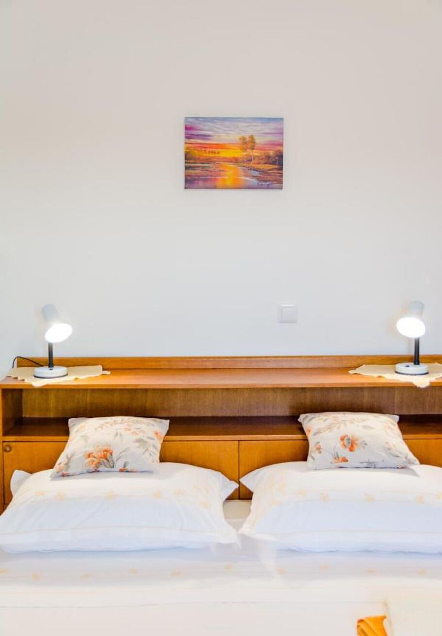 Ein Hotelzimmer mit Holz-Kopfteil, zwei Nachttischlampen, einem Sonnenuntergangsbild und Kissen mit Blumenmuster.