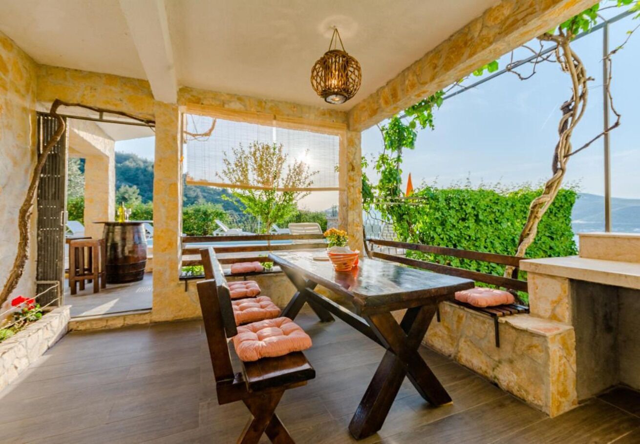 A dining area with a long table and benches, surrounded by stone walls and a view of greenery.