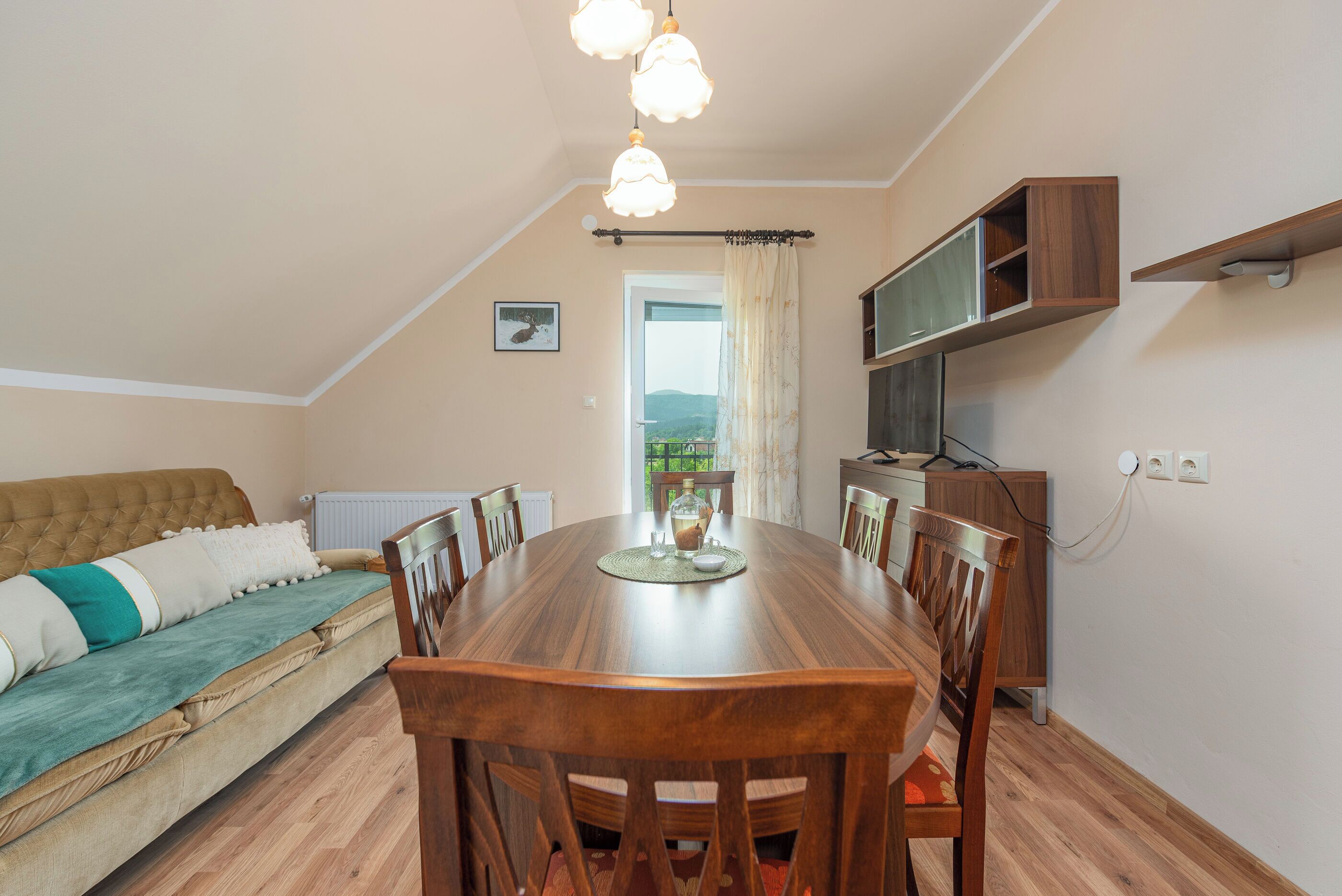 Un área de comedor con una mesa y sillas de madera, un sofá con una manta verde y vistas al exterior por una puerta.