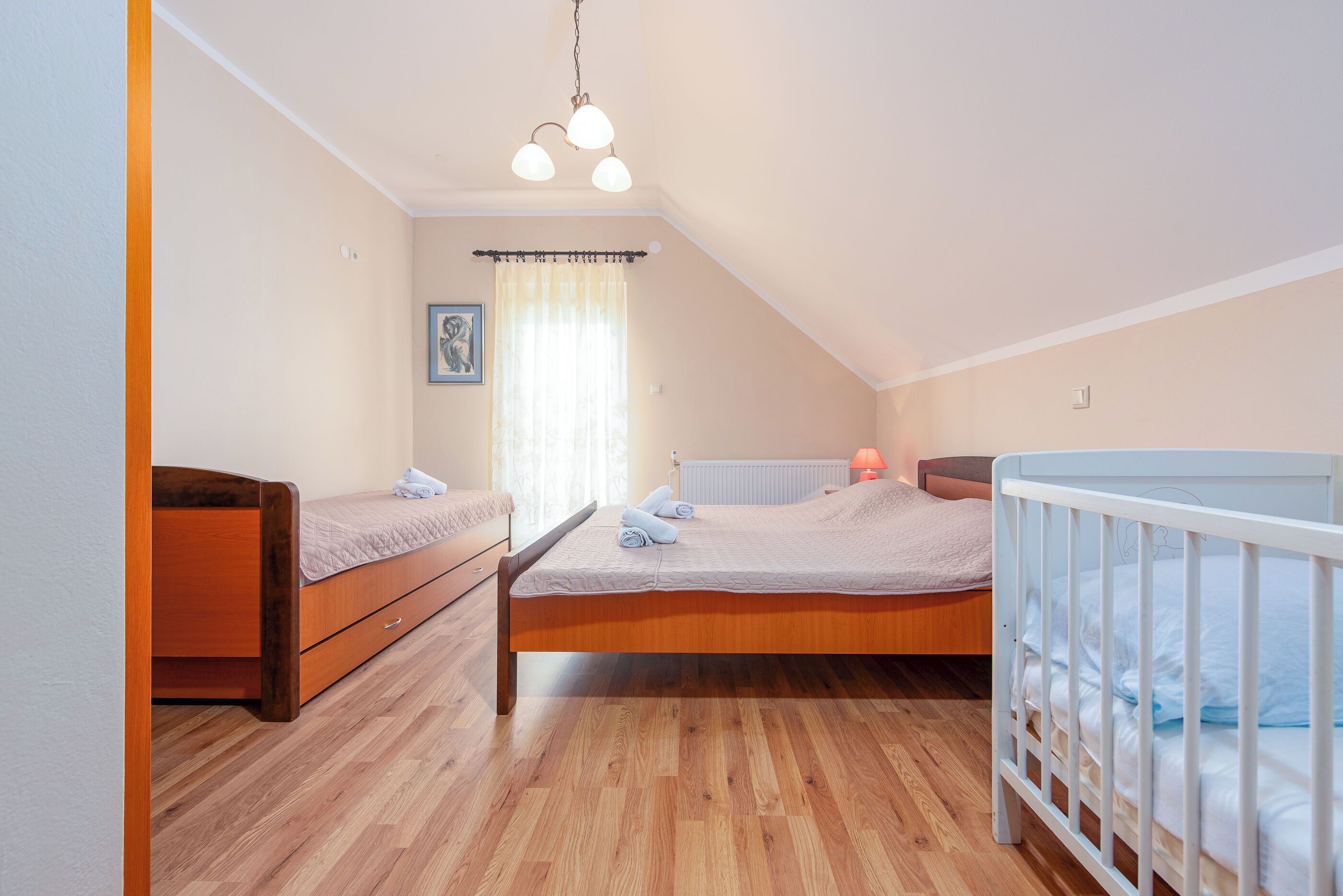 Een slaapkamer met een houten vloer, een bed met een wit hoofdeinde, een houten dressoir en een wit babybedje.