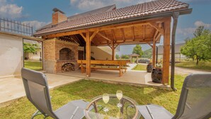 A covered outdoor seating area with a brick fireplace, a wooden pergola, and two lawn chairs with a glass table between them. - House Rose (Otocac)