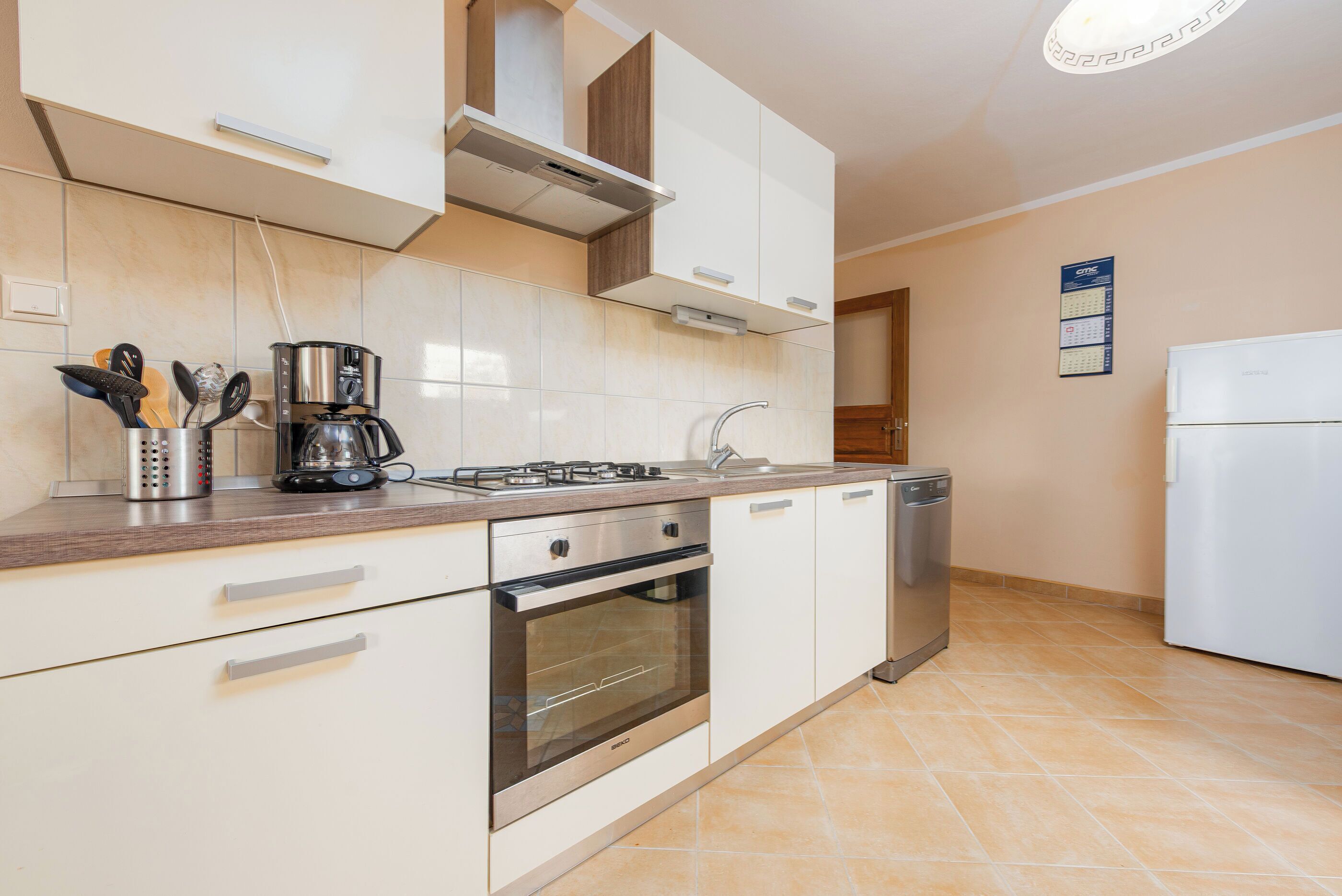 A modern kitchen with white cabinets, a stainless steel range hood, a built-in oven, a dishwasher, and a coffee maker.