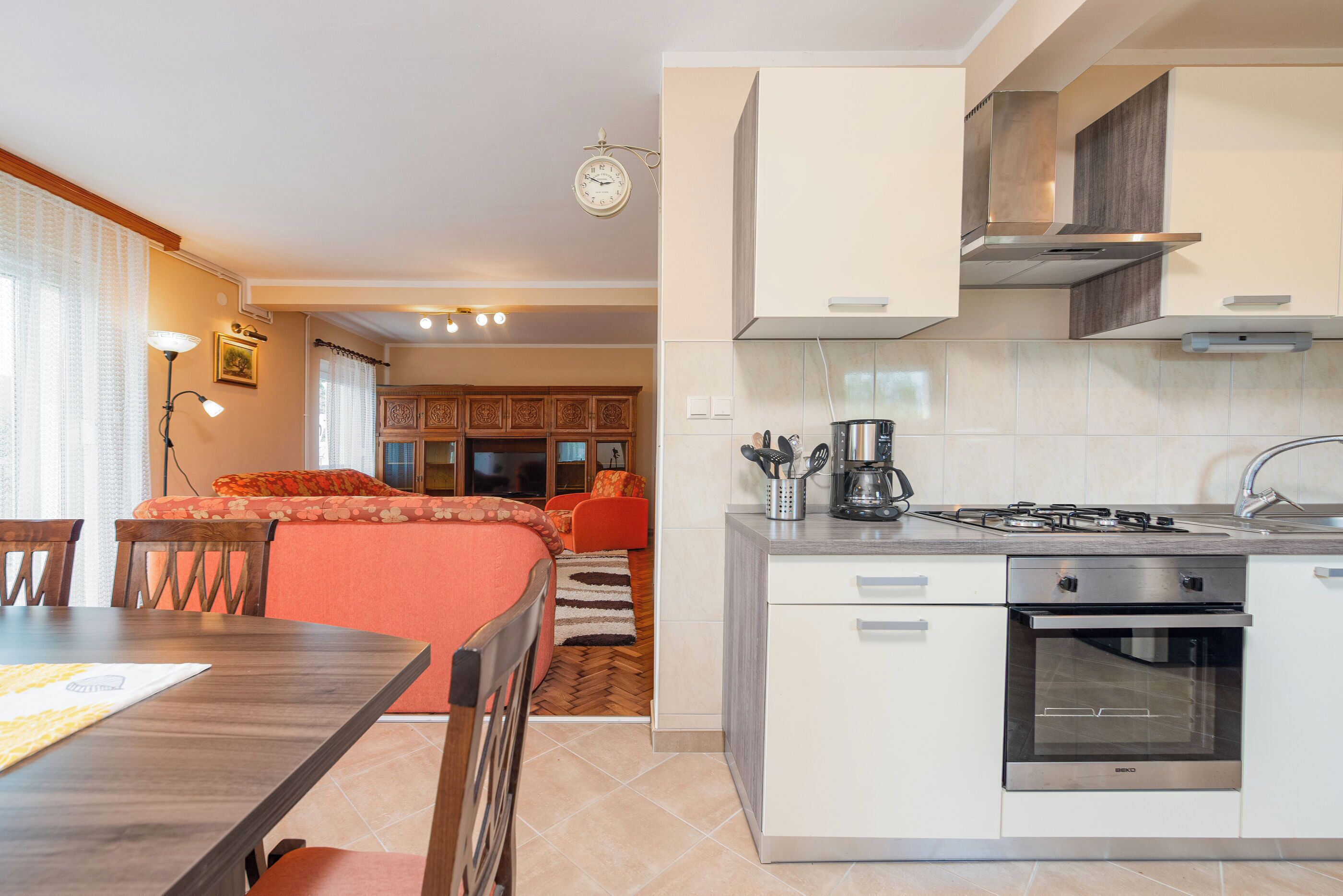 A modern kitchen with a built-in oven, a dining area with a wooden table and chairs, and a living area with a sofa and a coffee machine.