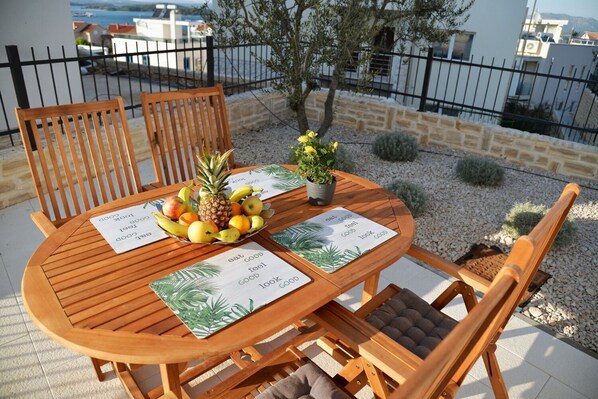 Een houten tafel met stoelen, een schaal met fruit en placemats waarop het woord 'good' herhaald is.