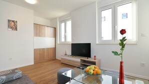 Egy modern nappali, melyben található egy síkképernyős televízió, egy gyümölcsökkel teli üveg kávézóasztal, egy szürke puff és egy vázában elhelyezett piros rózsa.