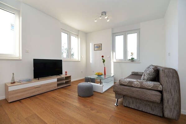 A modern living room with a flat-screen TV, wooden flooring, a grey sofa, and a glass coffee table. - Apartment Nata (Murter-Kornati)