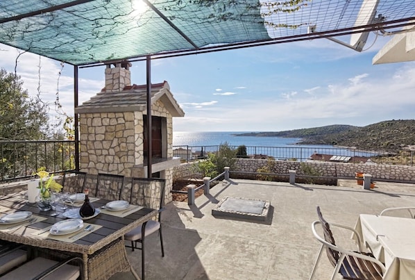 A covered outdoor dining area with a stone fireplace, a table set for two, and a scenic view of the sea and hills. - Apartments Apartmani Mastelic (Rogoznica)