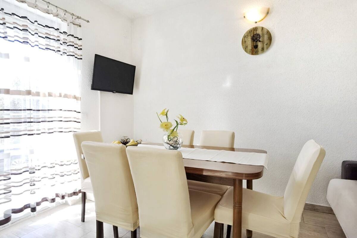 Un comedor con una mesa de madera, sillas color beige, un florero con flores, un televisor montado en la pared y un reloj.