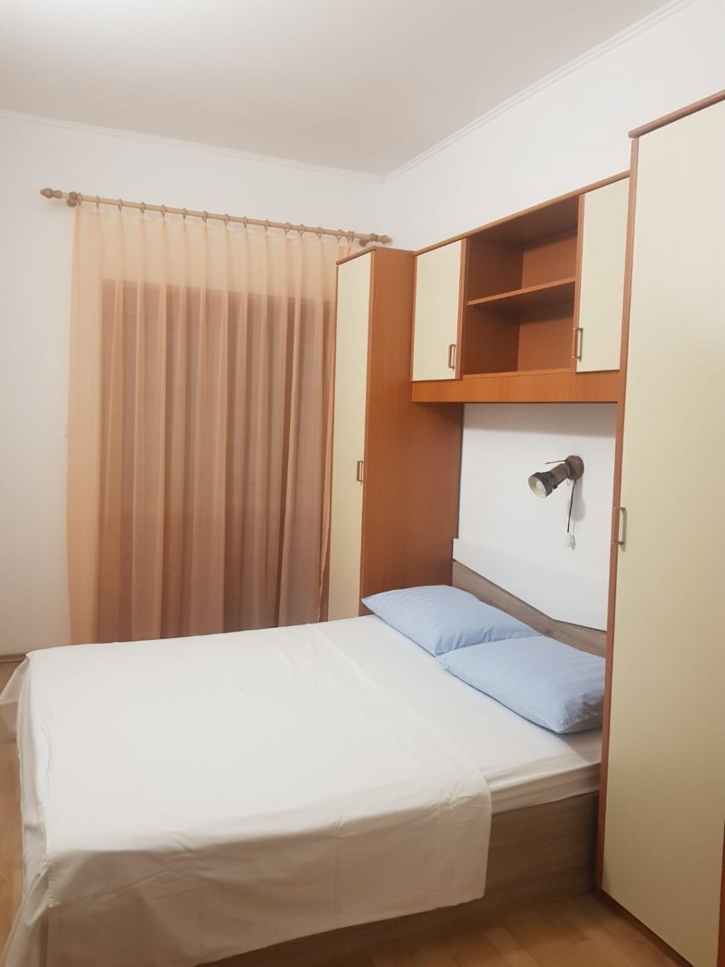 Une petite chambre avec un lit simple, une armoire en bois, une table de chevet et un rideau.
