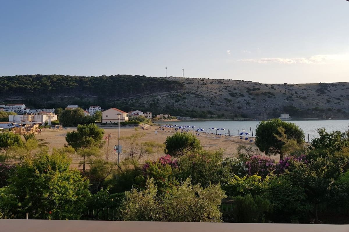 A coastal view with a beach, a hill with sparse vegetation, and buildings in the background.