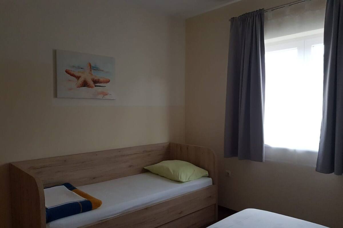 A small, simple room with a wooden bunk bed, a single bed, a window with curtains, and a painting on the wall.