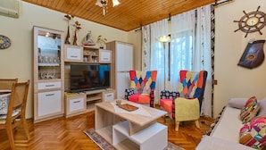 A cozy living room with a wooden ceiling, a television, a bookshelf, colorful chairs, and a patterned rug. - Apartment Giuseppe (Mali Losinj)