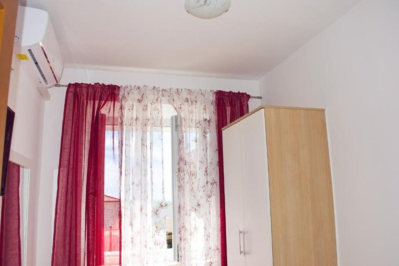 Et værelse med et træskab, et vindue med røde gardiner og en hvid aircondition.