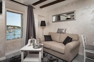 Ein gemütliches Zimmer mit einem beigen Sofa, einem kleinen Tisch mit Flasche und Gläsern, einer Lampe und Blick auf einen Hafen durch ein Fenster.