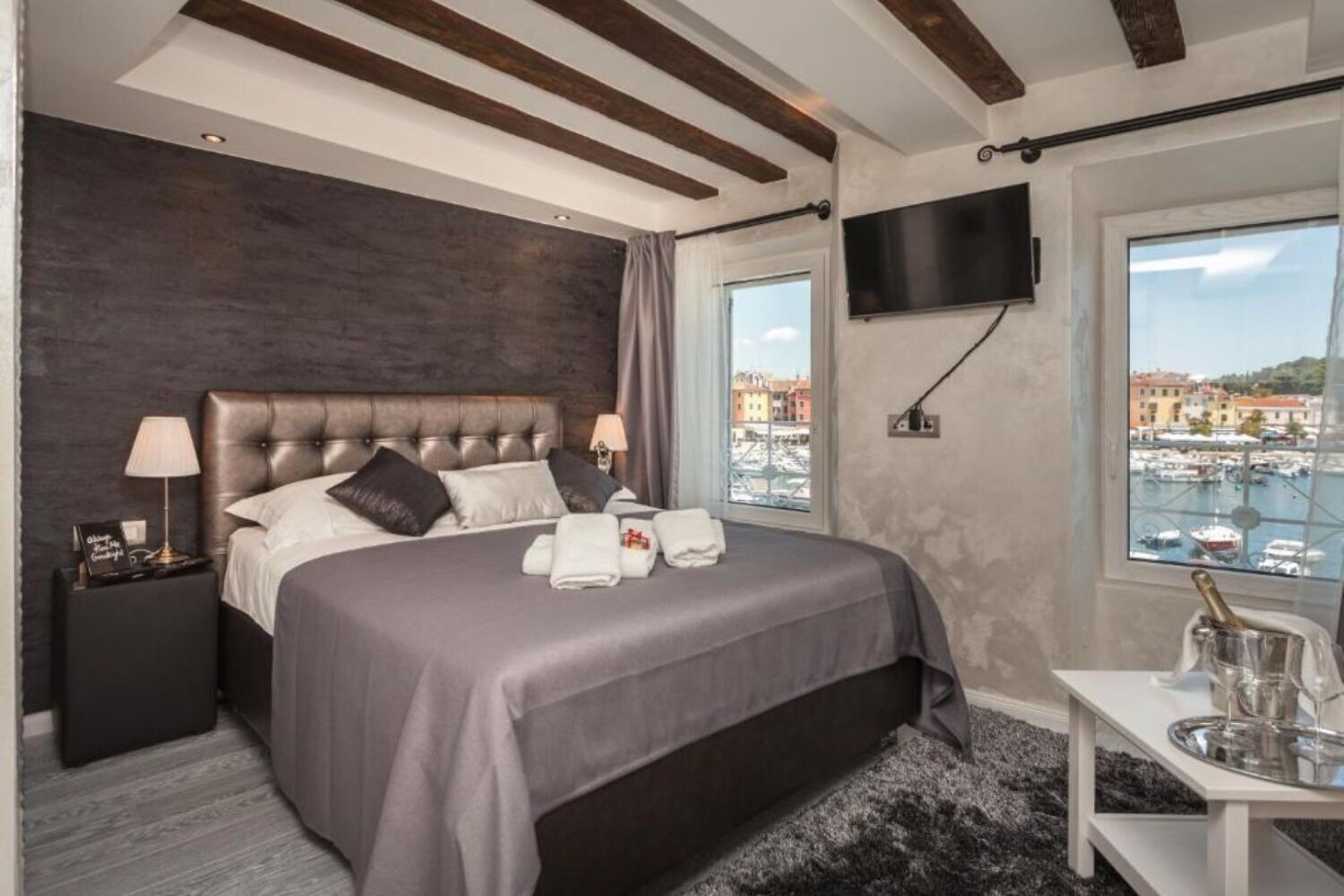 A modern hotel room with a large bed, a flat-screen TV, a view of the marina, and a small table with a bottle and glass.