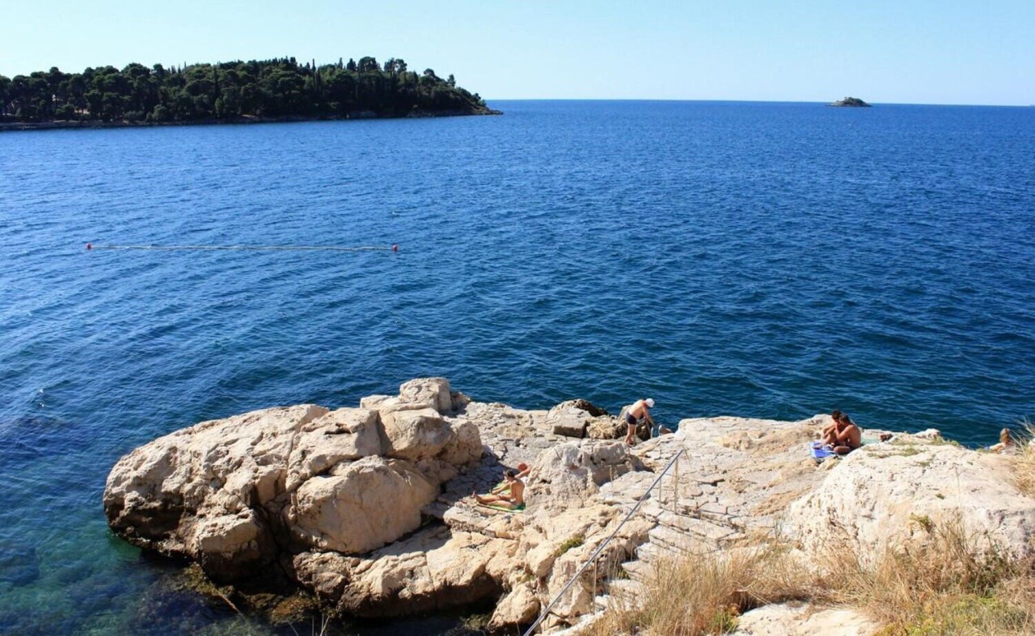 A rocky shoreline with clear blue sea, a distant island, and a few people enjoying the view.