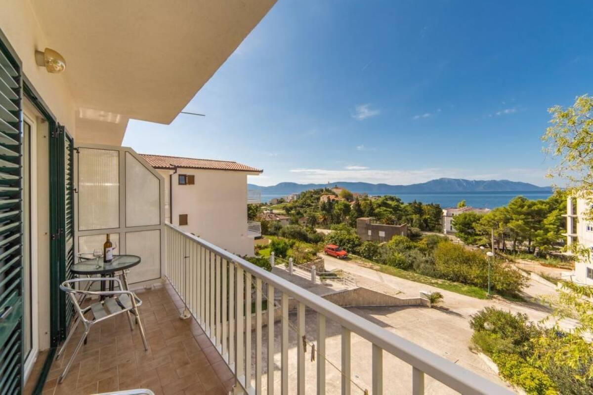 En balkon med et bord og stole, der giver udsigt over et boligområde med huse og et frit udsyn til havet og bjergene.