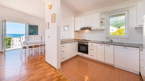 A modern kitchen with white cabinets, a stove, and a dishwasher. There is a dining area with a table and chairs, and a balcony with a view of the sea. - Apartments Gloria (Gradac)