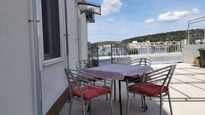 A covered outdoor patio with a table and chairs, a window, and a view of a body of water and buildings in the distance. - Apartment Tona (Tisno)
