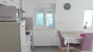 A compact kitchen with a microwave, a window with blinds, and a dining area with a table and chairs. - Apartment Tona (Tisno)