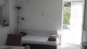 Quarto pequeno e moderno com cama, mesa e cadeira. Há uma janela com cortinas, um abajur e uma porta que dá para o exterior.