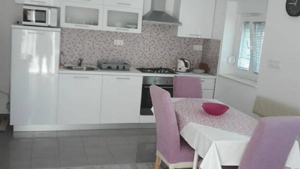 Cozinha compacta com armários brancos, micro-ondas, fogão e pia. Há uma mesa de jantar com cadeiras roxas e toalha de mesa branca.