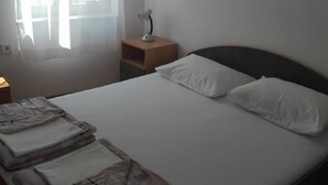 Uma cama bem arrumada com almofadas brancas e cobertores dobrados, uma mesa de cabeceira com abajur e uma janela com cortinas.