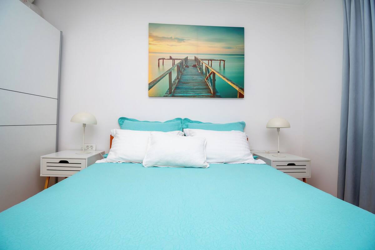 A bed with a teal bedspread, white pillows, and two bedside tables with lamps.