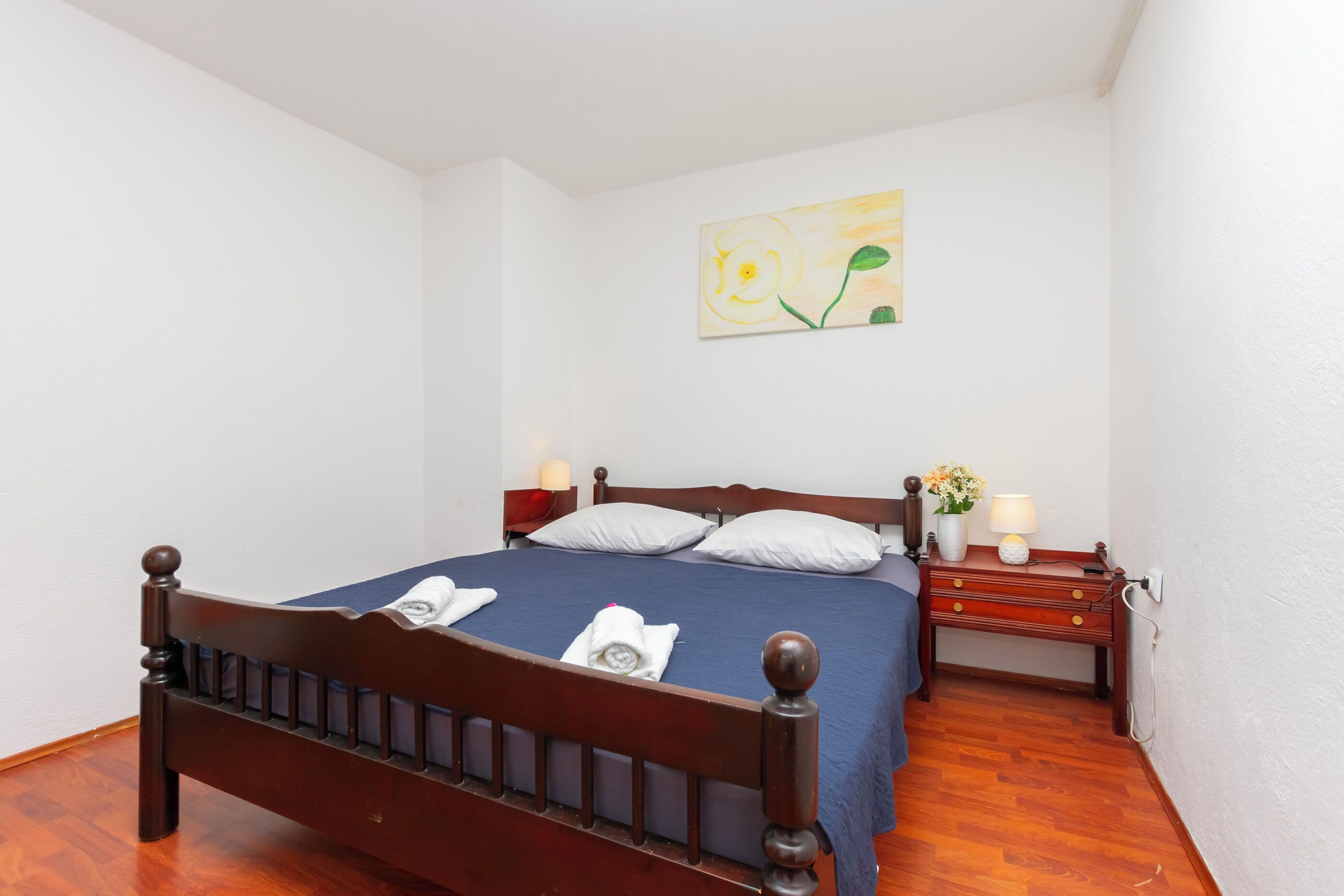 Un lit bien fait, avec une couvre-lit bleue et des oreillers blancs, une table de chevet en bois avec une lampe, et un tableau accroché au mur.