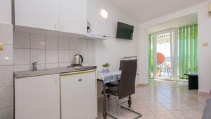 A compact kitchen with a built-in fridge, a kettle, and a small table with a chair. A TV is mounted on the wall, and a sliding door leads to an outdoor area with a table and chairs. - Apartments Zorica (Omis)