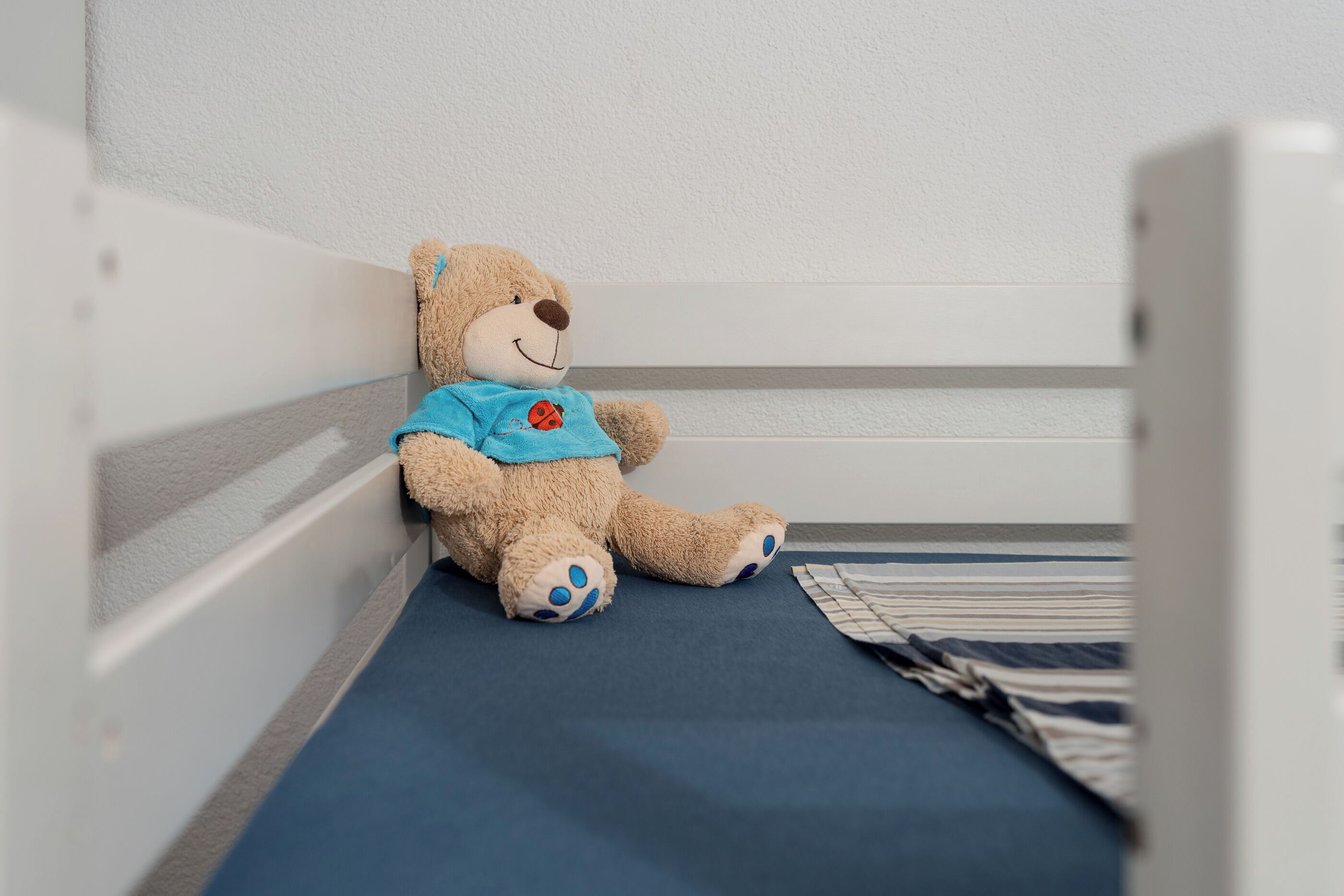 Nallekarhu istuu sinisen patjan päällä valkoisessa pinnasängyssä, jonka laidat ovat myös valkoiset.