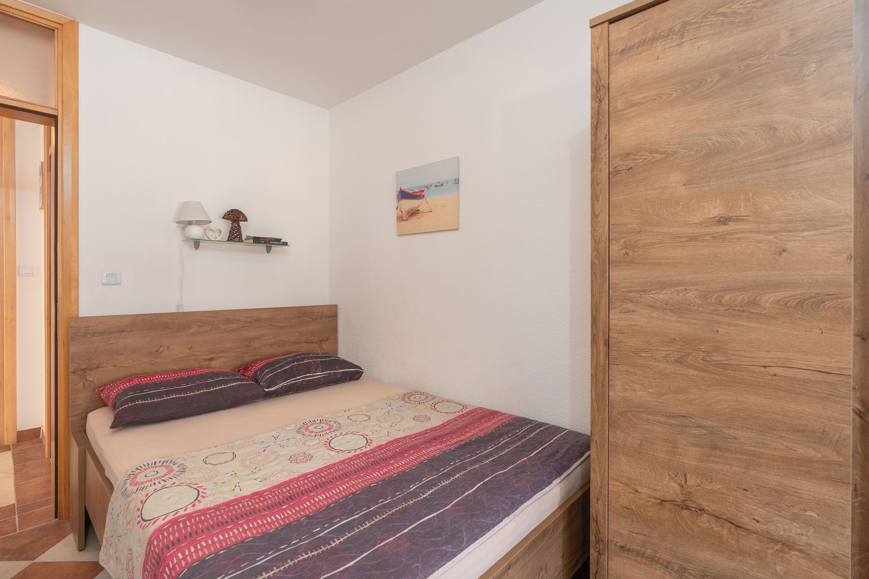A bedroom with a wooden headboard, a bed with patterned bedding, a small shelf with decorative items, and a beach-themed wall art.
