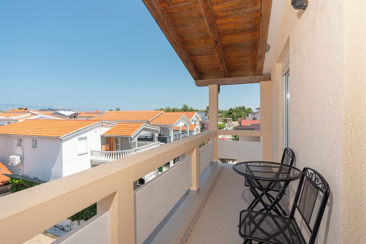Un balcone con soffitto in legno, un tavolo con sedie e vista sulle case vicine con tetti arancioni.