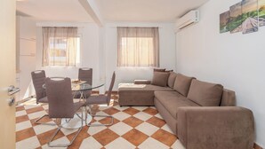 Ein modernes Wohnzimmer mit einem braunen Sofa, einem Glastisch und zwei Fenstern mit durchsichtigen Vorhängen.