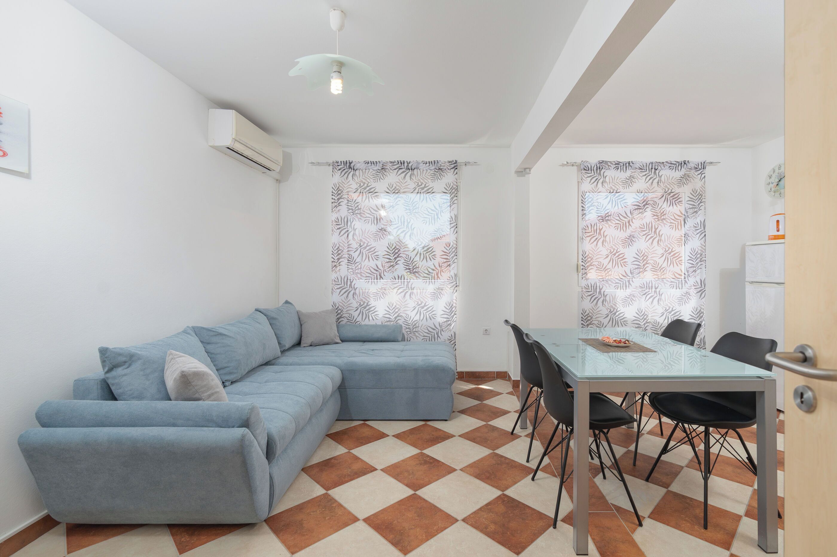 A modern living room with a blue sectional sofa, a glass dining table with four chairs, and patterned curtains on the windows.