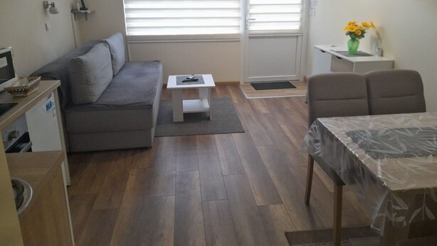 Ein kompakter Wohnraum mit grauem Sofa, kleinem weißen Tisch, Esstisch mit Plastikauflage, Wand-Fernseher und Fenster mit Jalousien.