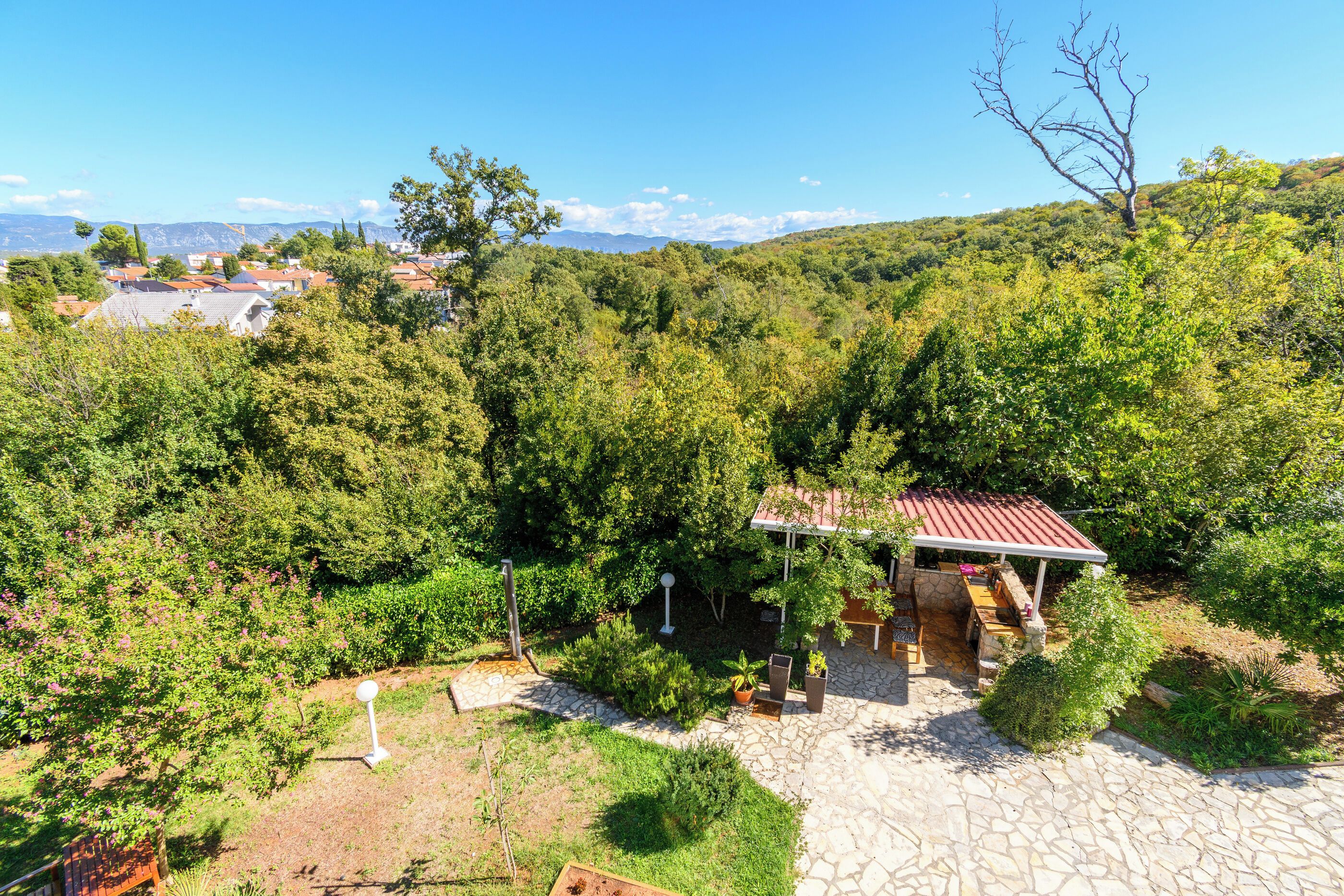 Un patio de piedra con pérgola, rodeado de abundante vegetación y vistas a un pueblo en la ladera.