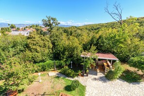 A stone patio with a pergola, surrounded by lush greenery and a view of a hillside village. - Apartments Vlado (Omisalj)