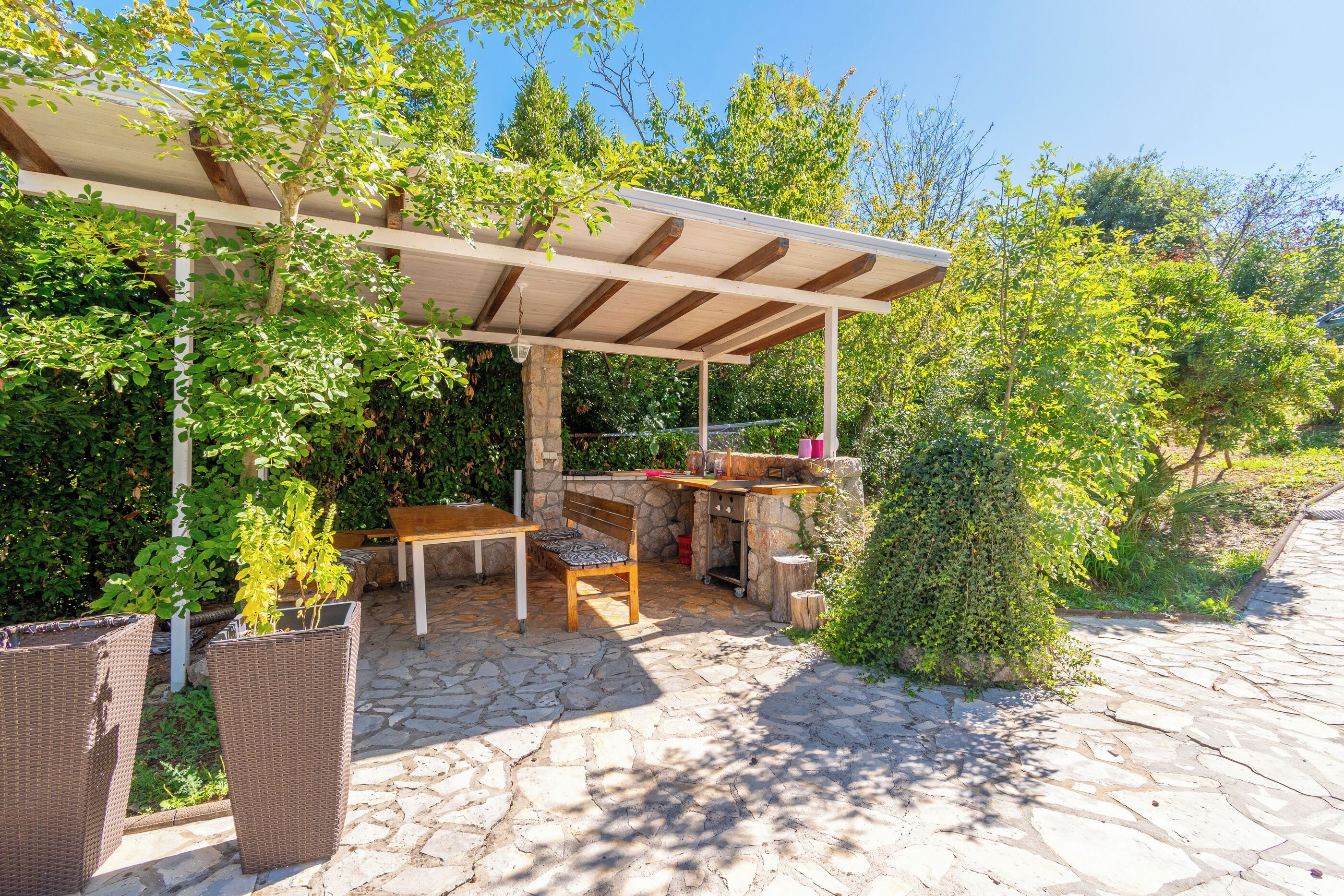 Eine Terrasse im Freien mit einer Pergola, Steinboden, einem Tisch, Stühlen und einer kleinen Küchenzeile.